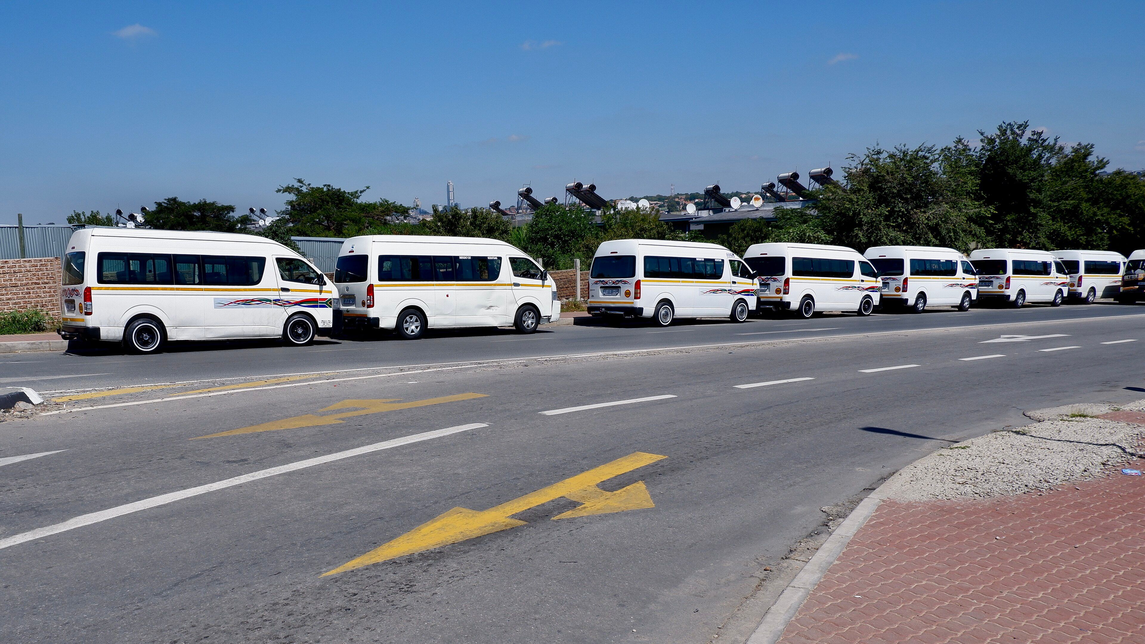 A row of white minibus taxis parked along a road in Alexandra, Johannesburg, with rooftops, solar geysers and the Sandton skyline behind