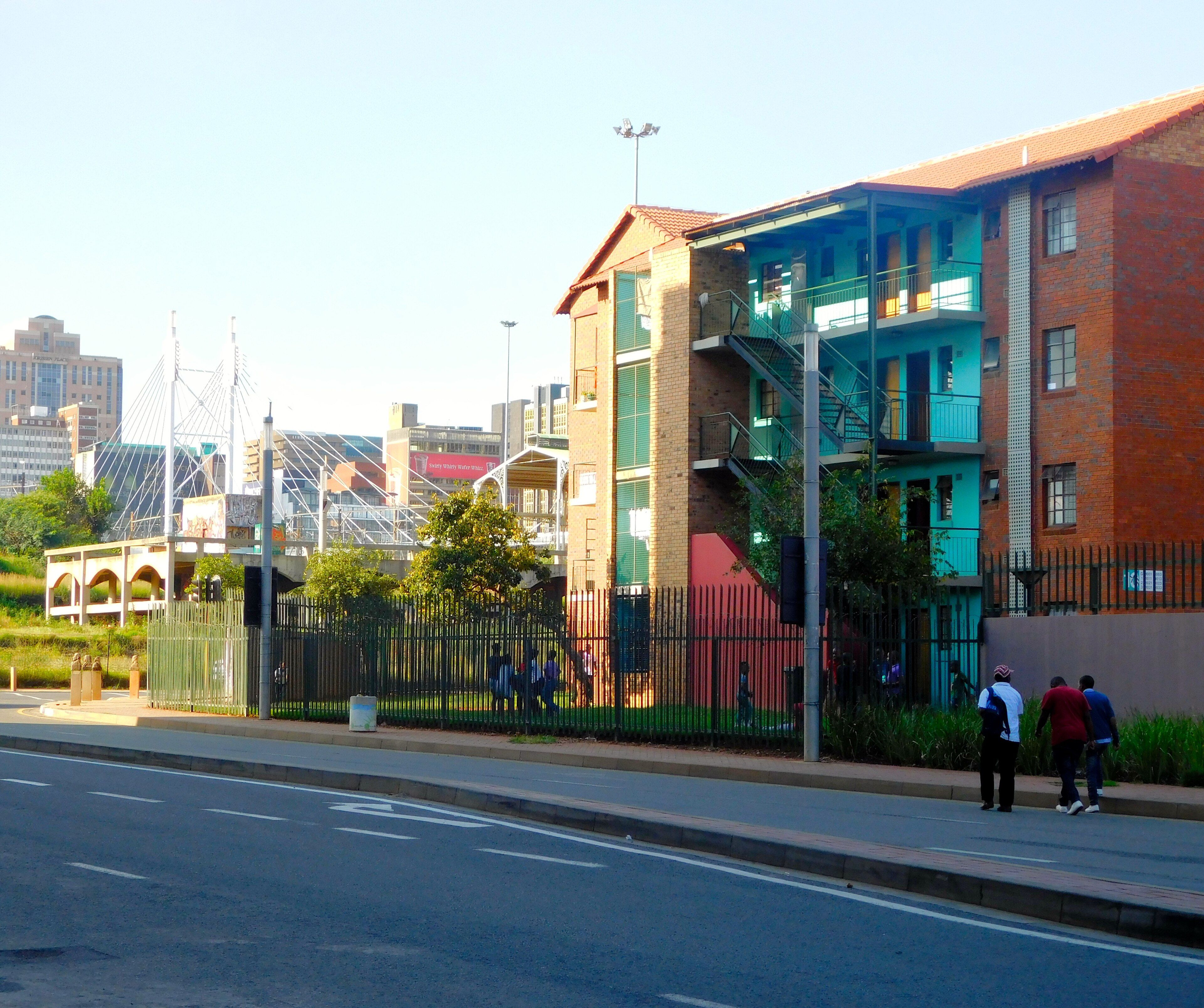 Three-storey brick walk-up rental flats with turquoise balconies and external stairs in Newtown, Johannesburg, with the Nelson Mandela Bridge and city buildings behind